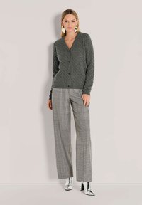 Frau, die eine graue Zopfstrickjacke, graue karierte weit geschnittene Hose, silberne Knöchelstiefel und große geometrische Ohrringe trägt, während sie vor einer hellen Wand steht.