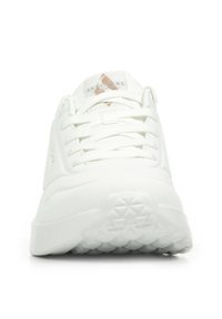 Vista frontale di una sneaker bianca con suola texture, lacci bianchi e un logo in tonalità oro e grigio sulla linguetta.