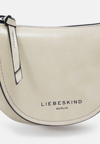 Beżowa skórzana torba na ramię o zaokrąglonym kształcie, z czarnymi szwami i tłoczonym logo "LIEBESKIND BERLIN" na dole. W zestawie regulowany pasek.