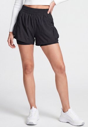 Kvinde iført sorte lagdelt atletikshorts, en hvid cropped langærmet top og hvide sneakers, står med hånden på hoften.