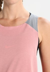 Lyserød sports tanktop med grå og sorte skulderdetaljer, lavet af blødt stof, med rund udskæring og diskret Nike-logo.