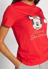 Žena v červeném vánočním tričku s Myškou Minnie v santovské čepici a nápisem "Veselé Vánoce", doplněná pruhovanými kalhotami.