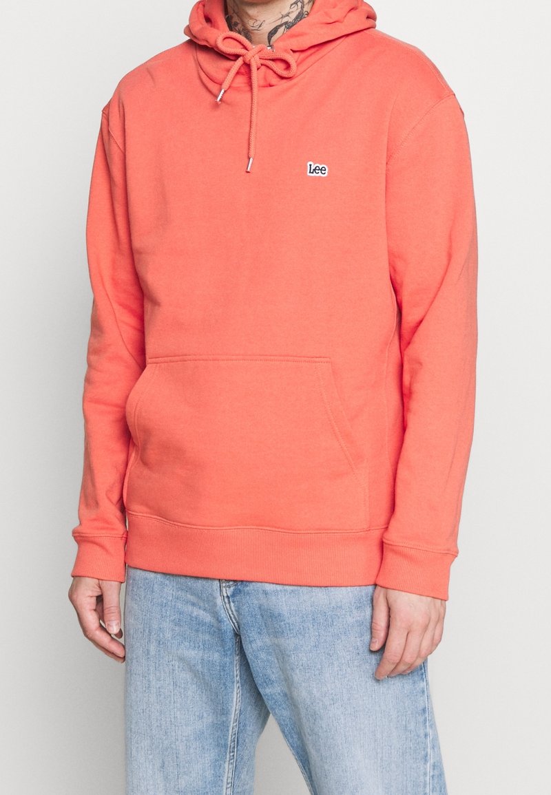Personne portant un sweat à capuche rose corail avec poche avant et logo "Lee" sur la poitrine, associé à un jean en denim bleu clair.