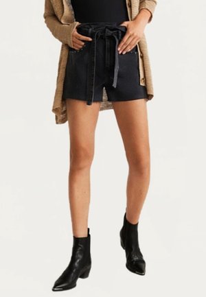 Mujer con pantalones cortos negros de cintura paperbag con cordón, cárdigan marrón claro, camiseta negra y botines negros, de pie con las manos cerca de los bolsillos de los pantalones cortos.