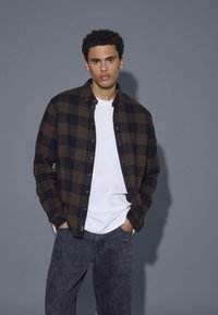 Rjav in črn karirast flanelasti srajc čez belo majico, v kombinaciji s temno sivimi kavbojkami. Model stoji z rokami v žepih ob sivi ozadju.