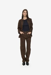 Donna in piedi con le mani in tasca, veste una giacca marrone con motivo e pantaloni abbinati, una maglietta nera e scarpe nere con punta, su sfondo bianco.