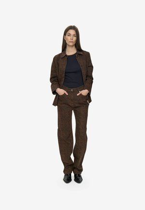 Donna in piedi con le mani in tasca, veste una giacca marrone con motivo e pantaloni abbinati, una maglietta nera e scarpe nere con punta, su sfondo bianco.
