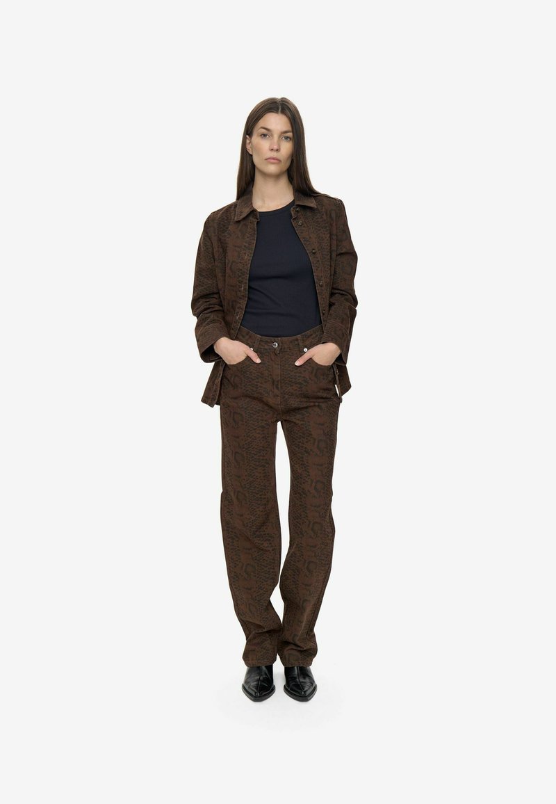 Donna in piedi con le mani in tasca, veste una giacca marrone con motivo e pantaloni abbinati, una maglietta nera e scarpe nere con punta, su sfondo bianco.