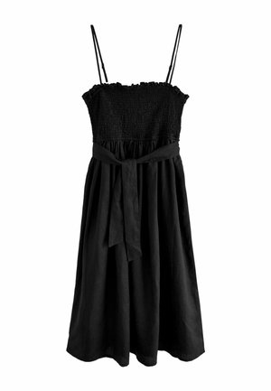 Femme portant une robe midi noire sans manches, des sandales plates noires et des bracelets argentés et dorés, debout à l'intérieur près d'une porte en bois et d'un canapé beige.