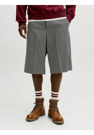 Fiú, szürke kockás térdnadrágot, csíkos zoknit, barna fűzős csizmát és bordó pulóvert visel, fehér háttéren áll.