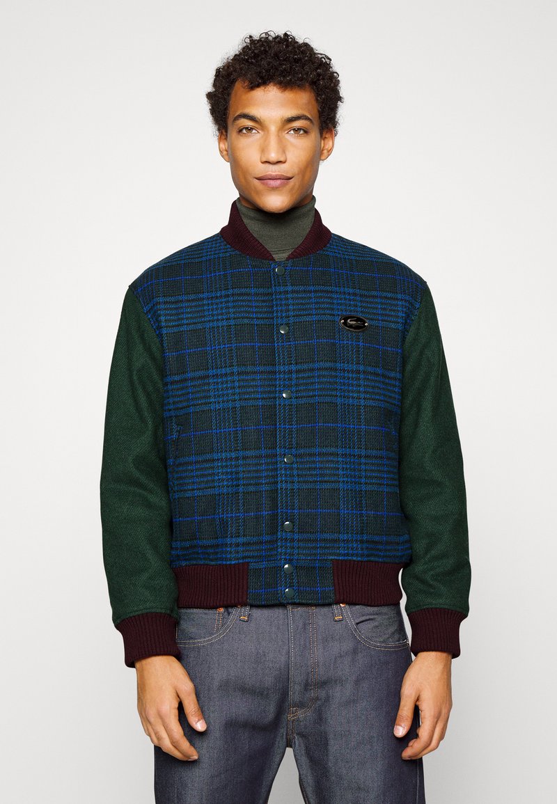 Homme portant une veste boutonnée en vichy bleu et vert foncé avec un col et des poignets bordeaux, associée à un jean noir, devant un mur uni.