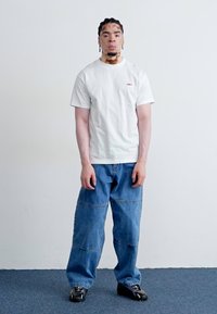 T-shirt branca de manga curta com logótipo vermelho, combinada com jeans azuis de corte largo e sapatos pretos. Fundo simples, sujeito em pé.