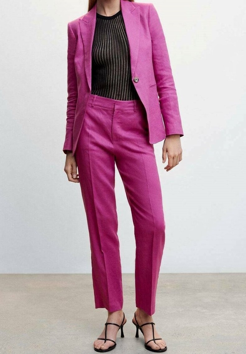Femme portant un tailleur rose vif avec une chemise rayée noire et marron et des sandales à talons hauts noires à brides sur fond uni.