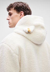 Jachetă albă din fâș cu o textură pufoasă, glugă supradimensionată și un mic eticheta galbenă atașată. Material neted și design lejer.