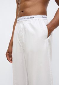 Λευκό βαμβακερό παντελόνι με ελαστική μέση που φέρει το γκρι και μαύρο λογότυπο "Calvin Klein". Πλαϊνές τσέπες και άνετη εφαρμογή.