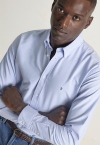 Camisa azul claro de manga larga, hecha de algodón, con un logo en el pecho y un cuello relajado. Se lleva con un cinturón trenzado.