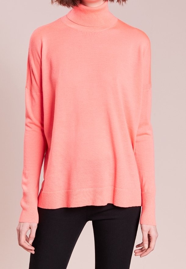 Femme portant un pull col roulé rose corail à manches longues associé à un pantalon noir, sur un fond neutre.