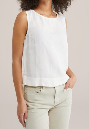 Femme aux cheveux bouclés portant un haut blanc sans manches et un pantalon beige clair, main dans la poche, fond neutre.