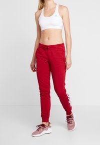Röda bomullsjoggers med elastisk midja och dragsko, med vit branding på vänster ben. Tillsammans med en vit sport-bh och stickade sneakers.
