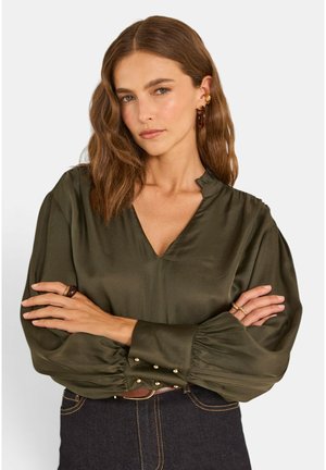 Femme aux cheveux bruns ondulés portant un chemisier vert foncé à manches longues avec des boutons dorés aux poignets et un jean foncé, les bras croisés.