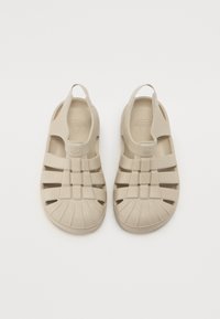 Beige műanyag szandál zárt orral, nyitott oldalakkal és hátsó sarokpánttal. Vízelvezetés céljából vízszintes és függőleges kivágásokkal rendelkezik.