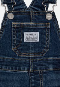 Tmavě modrý džínový overal s nastavitelnými kovovými přezkami, přední kapsou a kontrastním šitím. Na kapse je umístěna značená etiketa.
