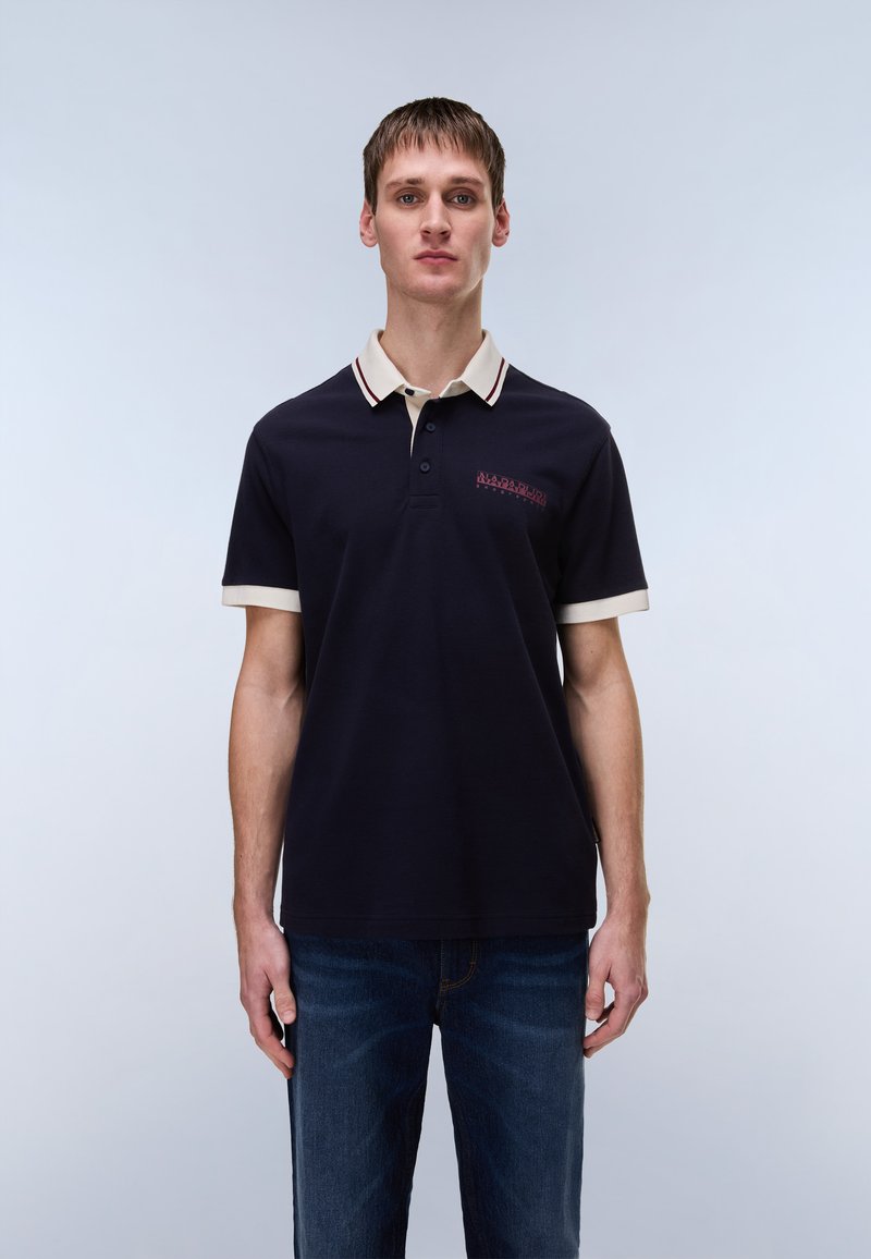 Μπλε σκούρο πόλο μπλουζάκι με μπεζ γιακά και κόκκινες λεπτομέρειες, με δύο κουμπιά και κεντημένο λογότυπο στο στήθος. Συνδυασμένο με σκούρα τζιν.