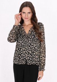 Blusa nera trasparente con stampa leopardata beige e scollatura arricciata. Maniche lunghe con polsini arricciati, tessuto leggero. Abbinata a pantaloni neri.