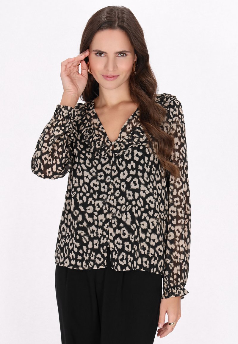Blusa nera trasparente con stampa leopardata beige e scollatura arricciata. Maniche lunghe con polsini arricciati, tessuto leggero. Abbinata a pantaloni neri.