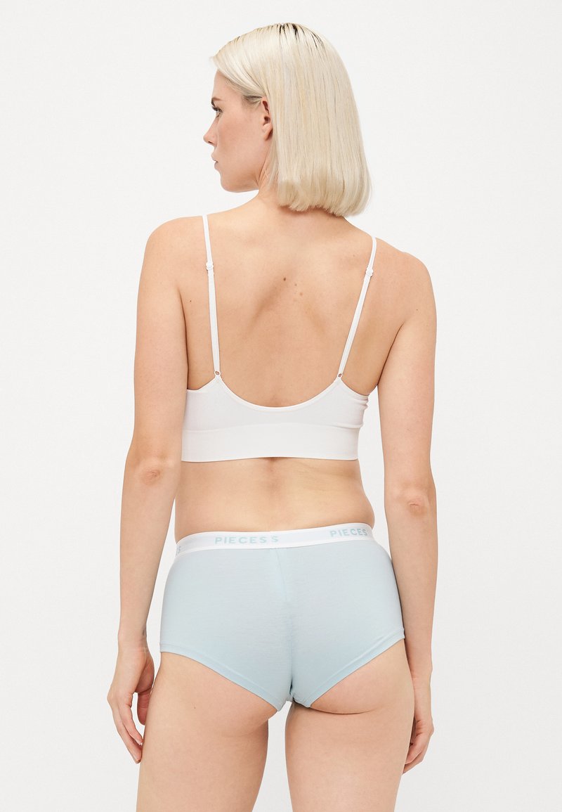 Kobieta z krótkimi blond włosami, nosząca białą braletkę i jasnoniebieskie majtki typu boyshort z napisem "PIECES" na pasku, pokazana od tyłu.