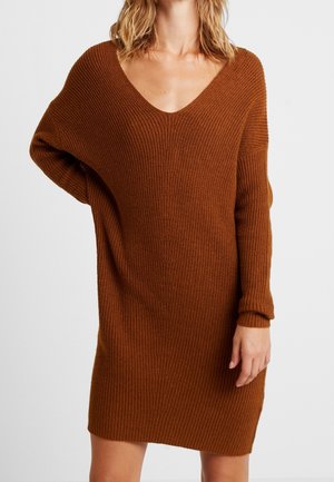 Femme portant une robe pull longue, ample et côtelée de couleur marron avec un décolleté en V profond et des manches longues, sur un fond clair uni.