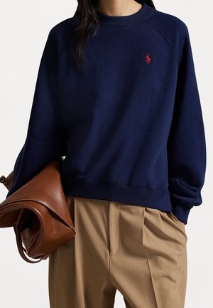 Femme portant un sweat-shirt bleu marine avec un logo rouge, un pantalon beige et tenant un sac à main plié en cuir marron, montrée du buste à la mi-cuisse.