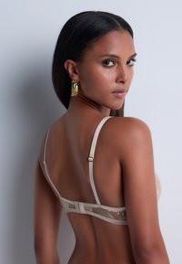 Femme aux cheveux bruns portant un soutien-gorge en dentelle beige et des boucles d'oreilles rectangulaires dorées se retourna contre un fond clair.