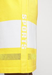 Keltaiset urheilushortsit, joissa on valkoinen vaakasuora raita. Alahelmassa on keltainen nauha, jossa lukee sana "SPORTS" valkoisella.