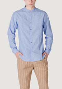 Camicia azzurro chiaro a maniche lunghe con colletto a band, patta dei bottoni e una trama sottile. Abbinata a pantaloni a righe color sabbia.