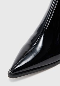 Sapato de salto alto em pele de verniz preta com bico fino. A superfície é brilhante com costura visível ao longo da borda. Sem adornos adicionais.