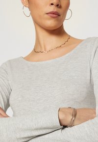 Graues Langarmshirt mit einem Bootsausschnitt, kombiniert mit einer wellenförmigen silbernen Halskette und drei dünnen silbernen Armreifen am Handgelenk.