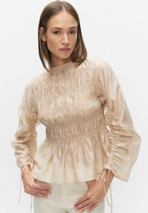 Kvinde med glat, brunt hår bærer beige langtærmet bluse med rynkede detaljer og lyse bukser, poserer med hænderne på hofterne.