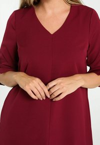 Femme portant une robe bordeaux à décolleté en V et manches aux coudes, les mains légèrement jointes au niveau de la taille.