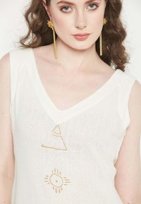 White linen top featuring a v-neckline and gold geometric patterns. Model wears long gold earrings; hair is styled in loose waves.