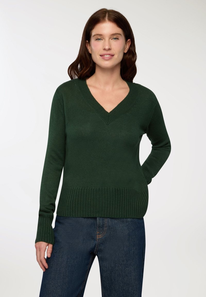 Ciemnozielony sweter z dekoltem w serek, z ryflowanym dołem i mankietami, o gładkiej fakturze i dopasowanym kroju, w połączeniu z niebieskimi dżinsami.