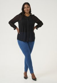 Sort langærmet bluse med struktureret overdel, gennemsigtig stof og knapdetalje; parret med blå skinny jeans og sorte spidse sko.