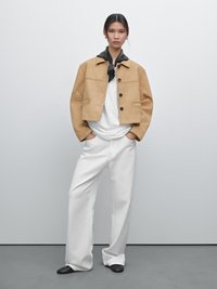 Tan cropped jasje met knopen aan de voorkant en twee zakken, gedragen over een witte T-shirt. Model draagt een witte broek en een zwarte sjaal.