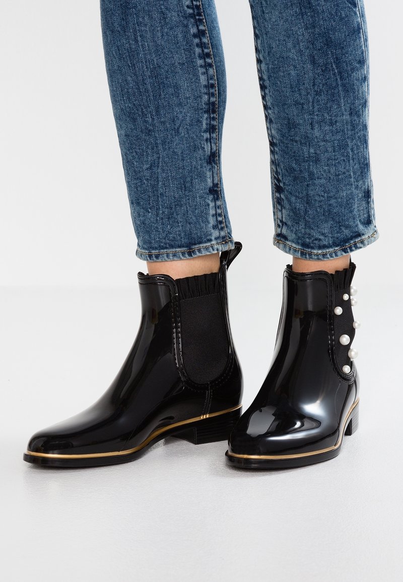 Bottines noires brillantes avec panneaux latéraux élastiques, détails dorés et accents de perles sur la botte droite. Portées avec un jean en denim raccourci.
