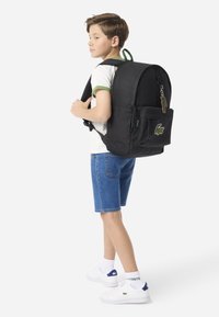 Schwarzer Rucksack mit strukturiertem Stoff, abgerundeter Form, vorderer Reißverschlusstasche und grünem Akzent. Verfügt über ein Lacoste-Logo und Schlüsselanhänger-Detail.