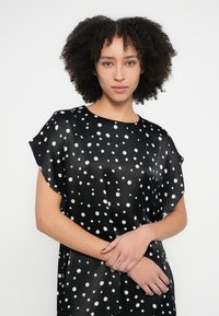 Schwarzes Satinkleid mit weißen Punkten, kurzen Ärmeln und lockerem Schnitt. Glatte Textur mit rundem Ausschnitt.