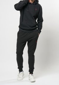 Svart huvtröja med dragsko och framficka, ihop med svarta sweatpants med en liten röd logotyp. Bärs med vita sneakers.