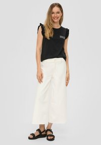 Schwarzes, kurzärmliges Oberteil mit Rüschelementen, weiße weite Hose und schwarze Riemchensandalen. Schlichte Gestaltung, weicher Stoff und lässiger Stil.