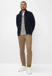 Marineblaue Strickjacke mit Knopfverschluss, kombiniert mit einem karierten Hemd, hellbraunen schmal geschnittenen Hosen und weißen Sneakers. Einfarbiger Hintergrund.