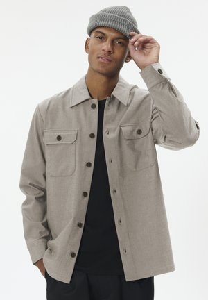Mann trägt eine hellbeige knöpfte Jacke, schwarzes Hemd, dunkle Hose und eine graue Strickmütze, berührt den Rand der Mütze.